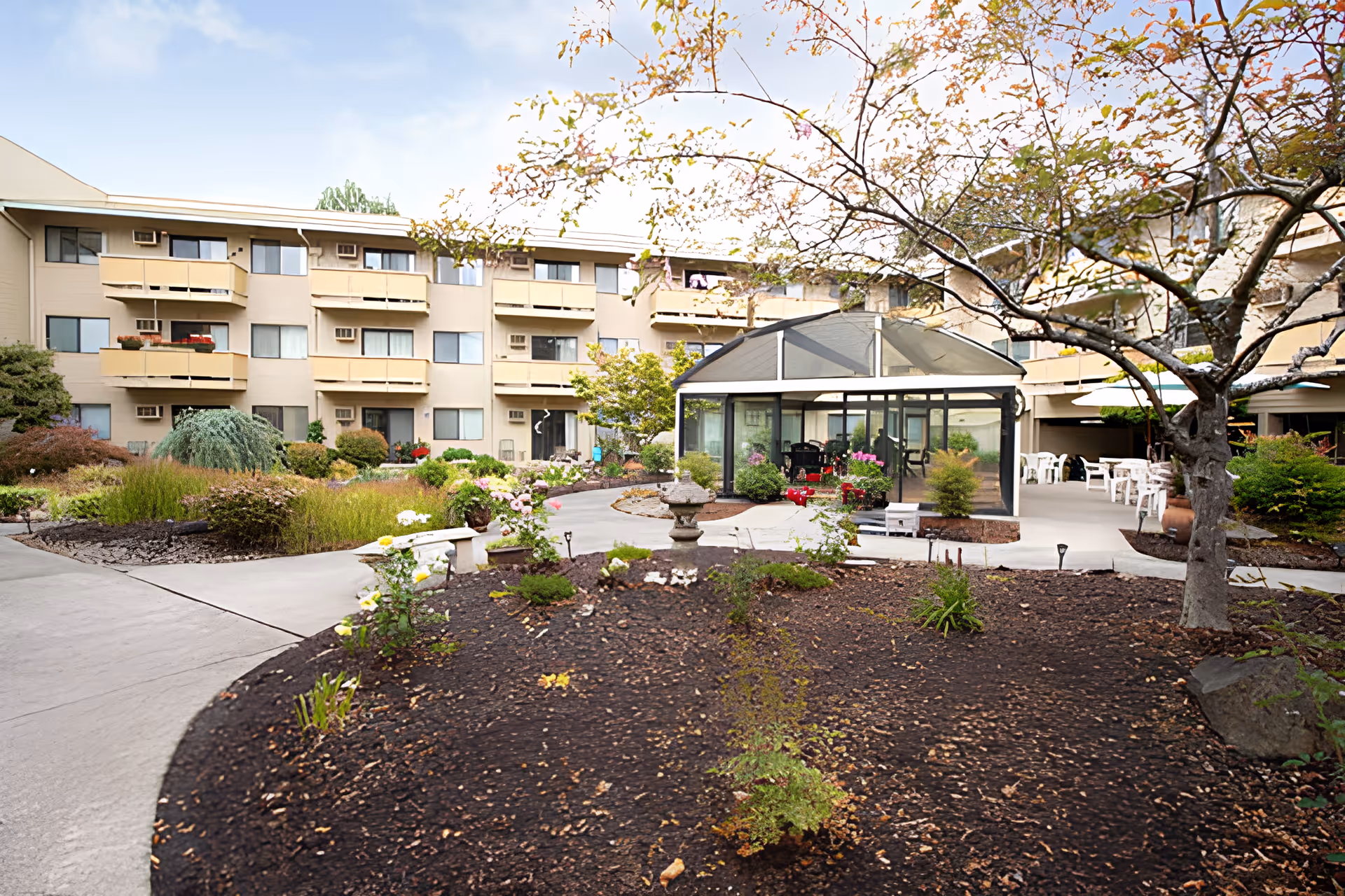 Courtyard of a senior living facility with landscaped beds, walkways, a glass-enclosed gazebo, and a multi-story building with balconies.
