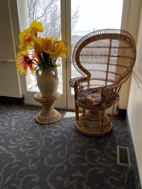 A wicker chair with a patterned cushion next to a small wicker table holding a large vase with bright yellow and orange flowers, positioned in front of a glass door letting in natural light.