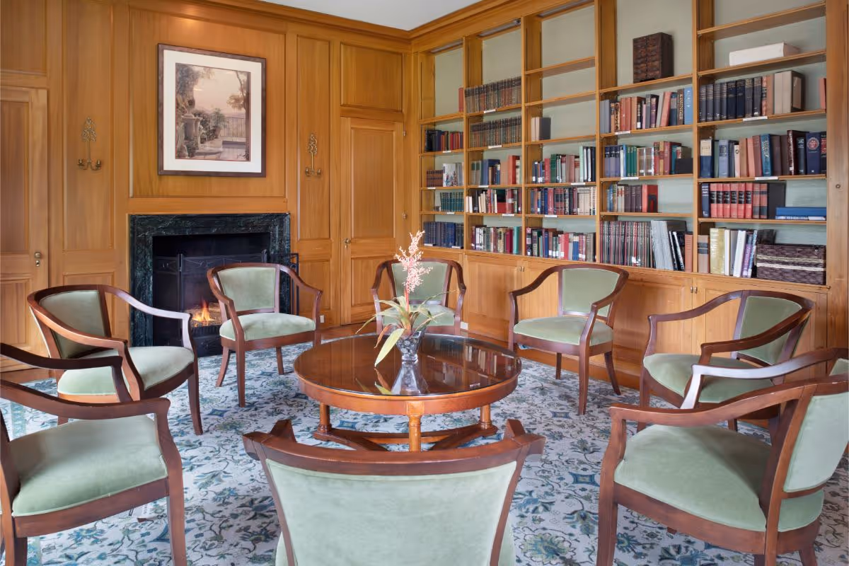 A cozy library room with wooden paneling and built-in bookshelves filled with books. There is a round wooden table with a small flower arrangement in the center, surrounded by seven wooden chairs with green cushions. A fireplace with a fire burning is set into the wood-paneled wall, above which hangs a framed picture of an outdoor scene.
