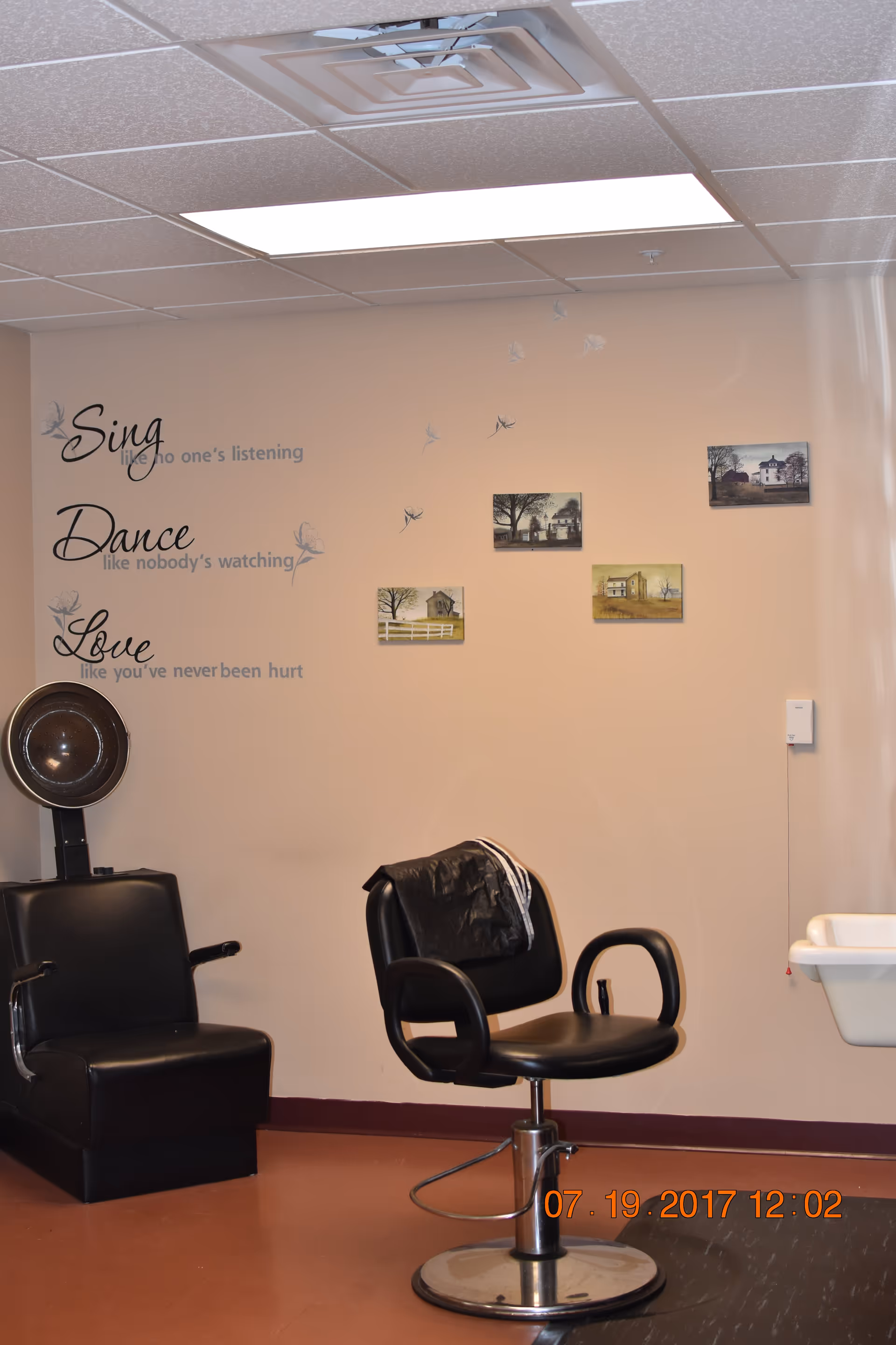Interior of a salon area in a senior living facility with a black salon chair and a hair dryer chair. The beige wall behind has decorative text that reads 'Sing like no one's listening, Dance like nobody's watching, Love like you've never been hurt' along with small framed pictures and dandelion decals. A white sink is visible on the right side.