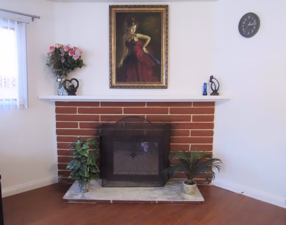 Brick fireplace with a white mantel topped by a framed portrait and decorative items, flanked by potted plants on a hardwood floor.