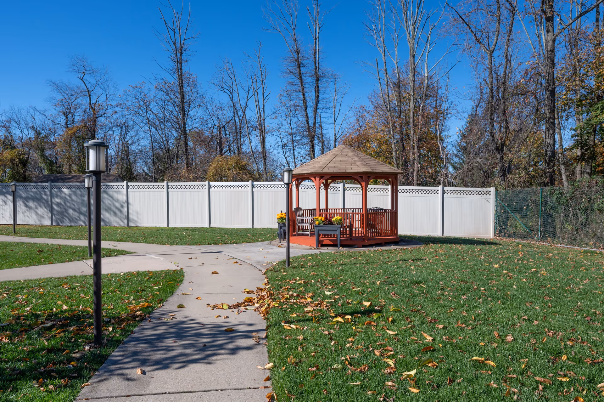 Outdoor area with a concrete pathway leading to a wooden gazebo surrounded by green grass and some fallen leaves. There are lamp posts along the pathway and a white fence in the background with leafless trees beyond it under a clear blue sky.