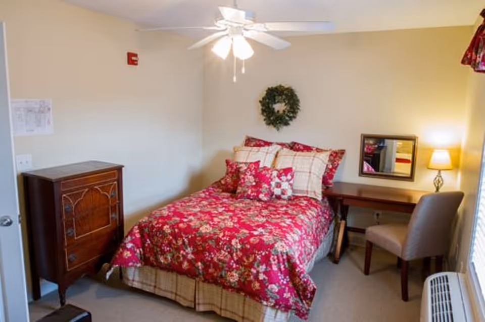 Small furnished bedroom with a red floral bedspread, wooden dresser, desk with chair and lamp, ceiling fan, and a wreath on the wall.