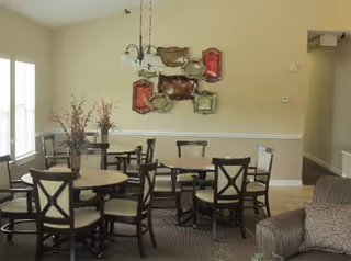 Well-lit communal dining area with several round tables and chairs, a chandelier, wall-mounted decorative art, and a nearby seating area.