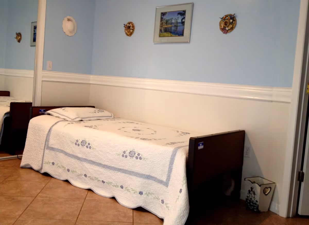 A small bedroom with a single bed covered in a white quilt with floral patterns. The walls are painted light blue on the upper half and white on the lower half, with decorative wall hangings and a framed picture. There is a mirrored closet door on the left side and a decorative wastebasket on the floor near the bed.