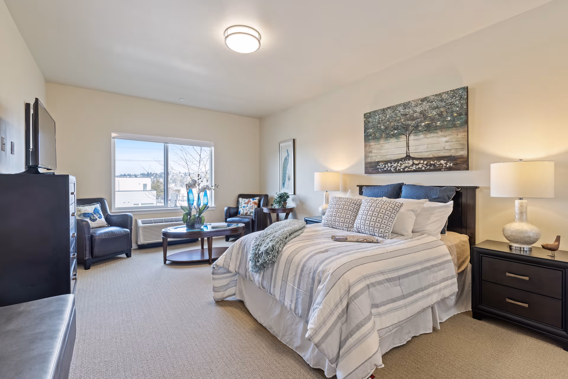 Well-lit furnished bedroom with a made bed, two nightstands and lamps, seating area with armchairs and a coffee table, and a window view.