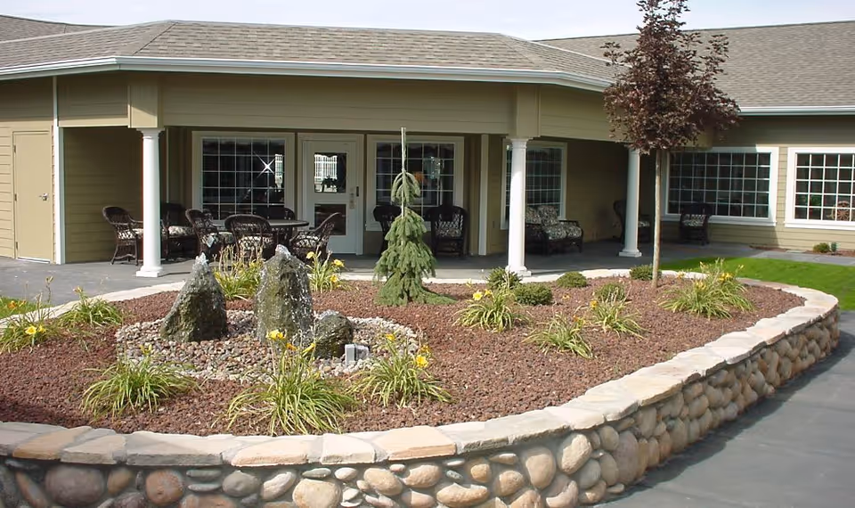 Outdoor garden area with a stone-bordered flower bed containing small plants, yellow flowers, and a small tree. Behind the garden is a covered patio with wicker chairs and tables, and large windows and doors of a building.