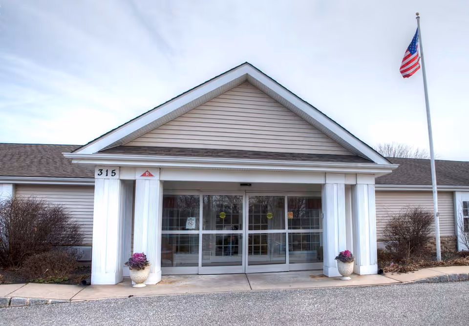 Front entrance of a single-story building with beige siding and a triangular roof overhang supported by white columns. There are two potted plants with purple flowers on either side of the automatic sliding glass doors. An American flag is flying on a flagpole to the right of the entrance. The building number 315 is displayed on the left column.
