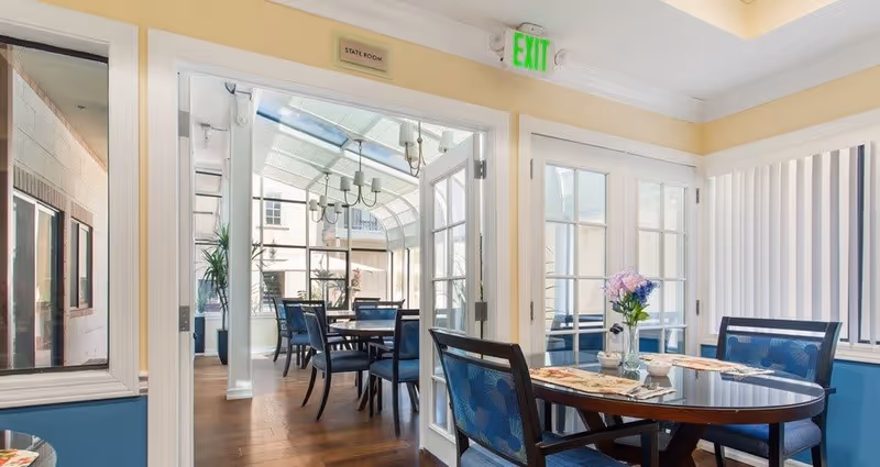 Interior view of a dining area in a senior living facility with round tables and blue cushioned chairs. The room has large windows and glass doors leading to a sunlit room with additional tables and chairs. A vase with flowers is placed on the table in the foreground. The walls are painted light yellow and blue, and there is an exit sign above the door.
