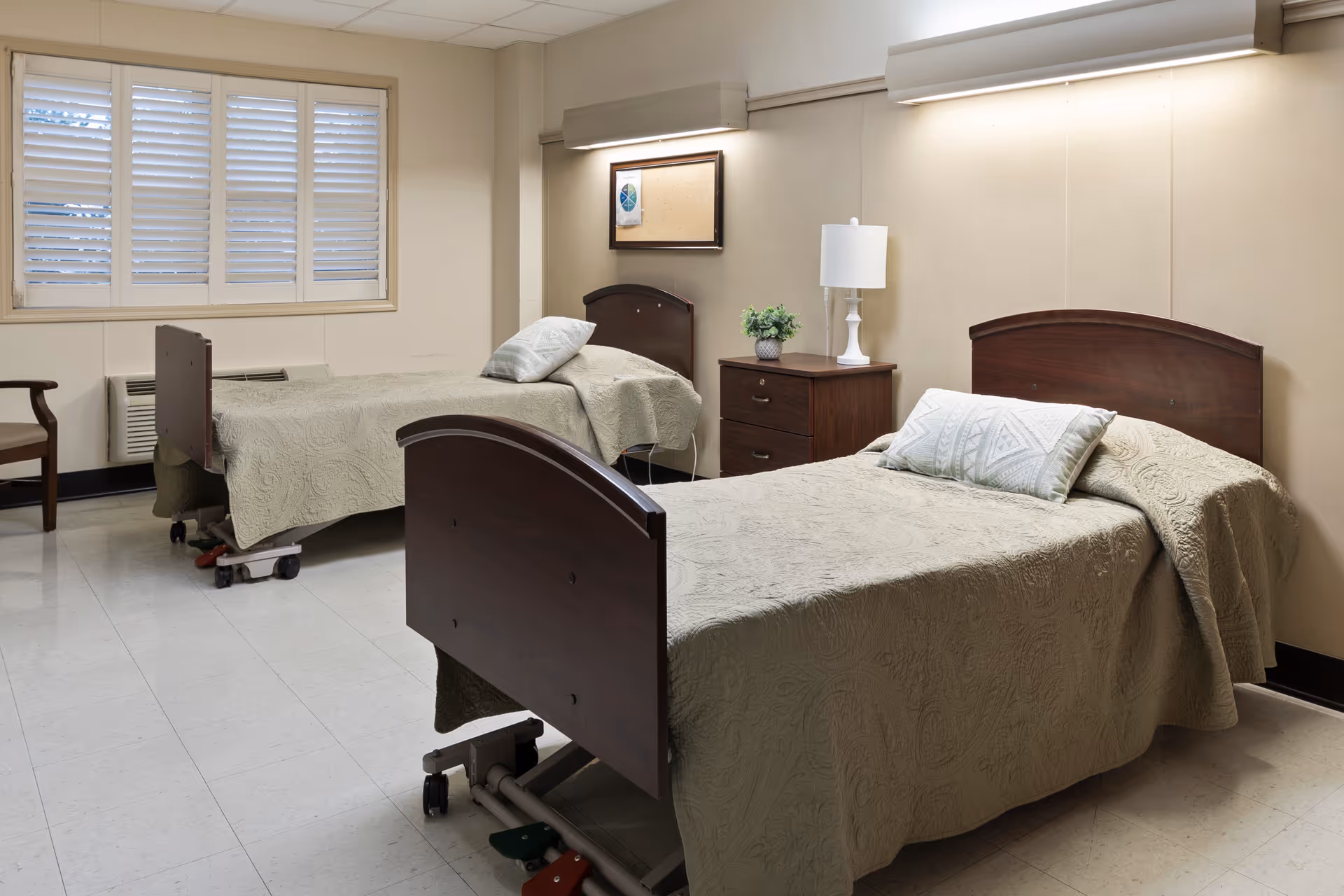A simple, clean room with two hospital-style beds covered in beige bedspreads, each with a pillow. Between the beds is a wooden nightstand with a white lamp and a small plant. The room has beige walls, a window with white shutters, and a chair near the window.