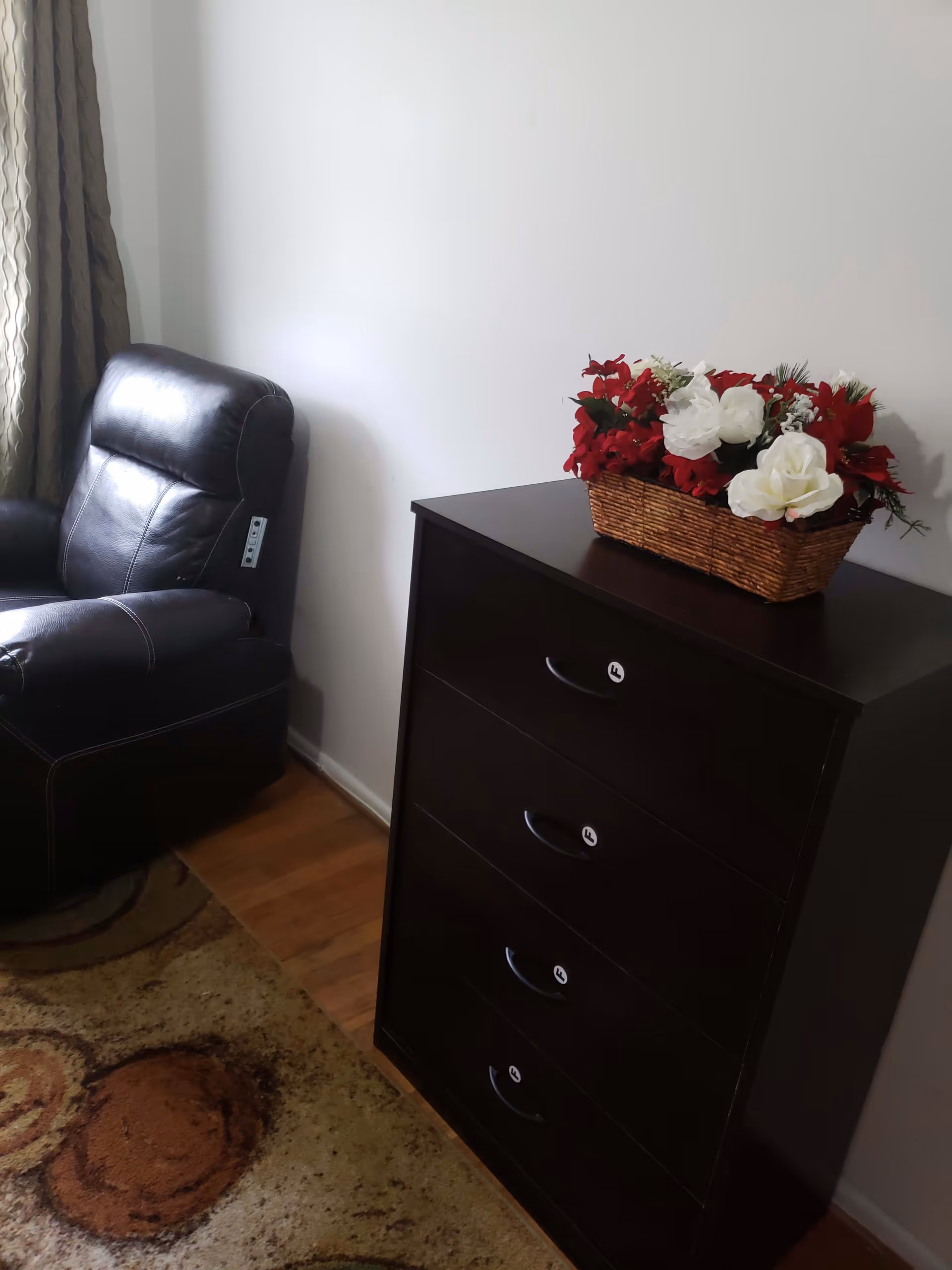 A corner of a room featuring a black leather recliner chair next to a dark wooden chest of drawers with a basket of red and white artificial flowers on top. The floor has a patterned carpet and there is a curtain partially visible on the left side.