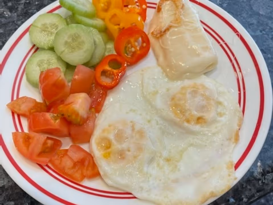 A plate with two fried eggs, sliced cucumbers, chopped tomatoes, sliced red and yellow bell peppers, and a piece of cooked white fish or chicken on a white plate with red stripes.