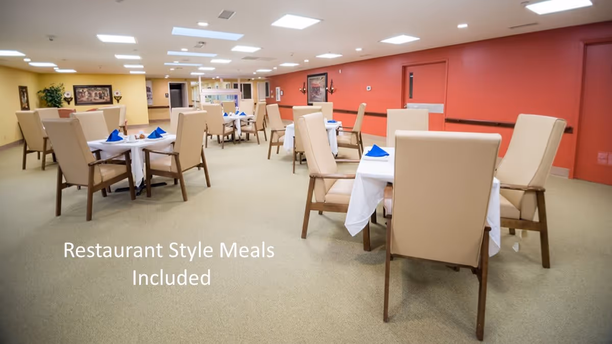 A dining room with multiple tables covered with white tablecloths and set with blue napkins. The room has beige cushioned chairs, a carpeted floor, and walls painted in yellow and red tones. There are framed pictures and plants decorating the walls.