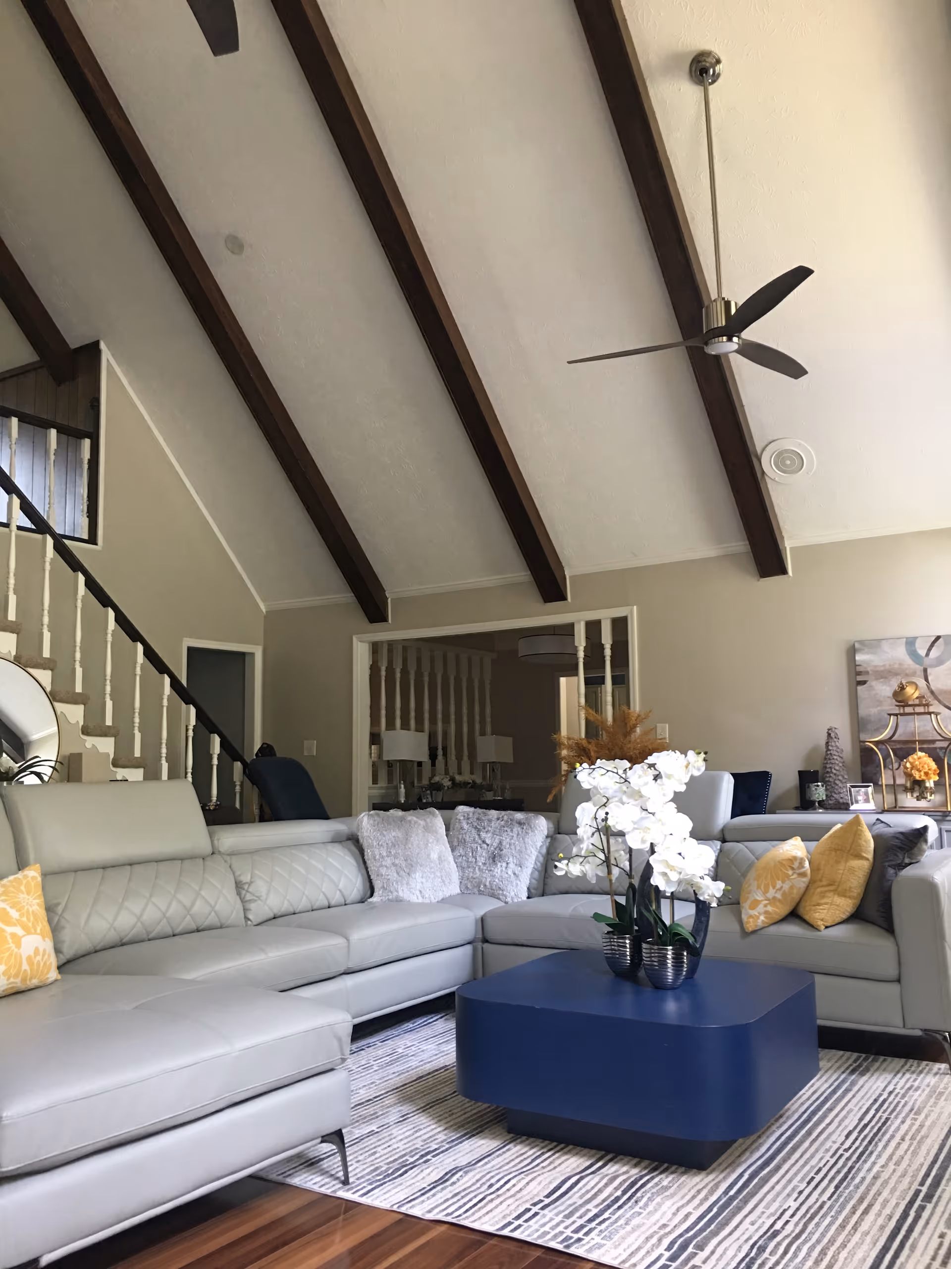 Spacious living room with vaulted ceiling and exposed wooden beams, a gray sectional sofa, blue coffee table and a white orchid centerpiece.