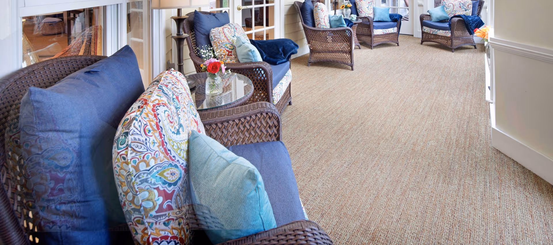 A cozy indoor seating area with wicker armchairs featuring blue and patterned cushions, small glass-top side tables with flower vases, beige carpeted floor, and light-colored walls with white trim.