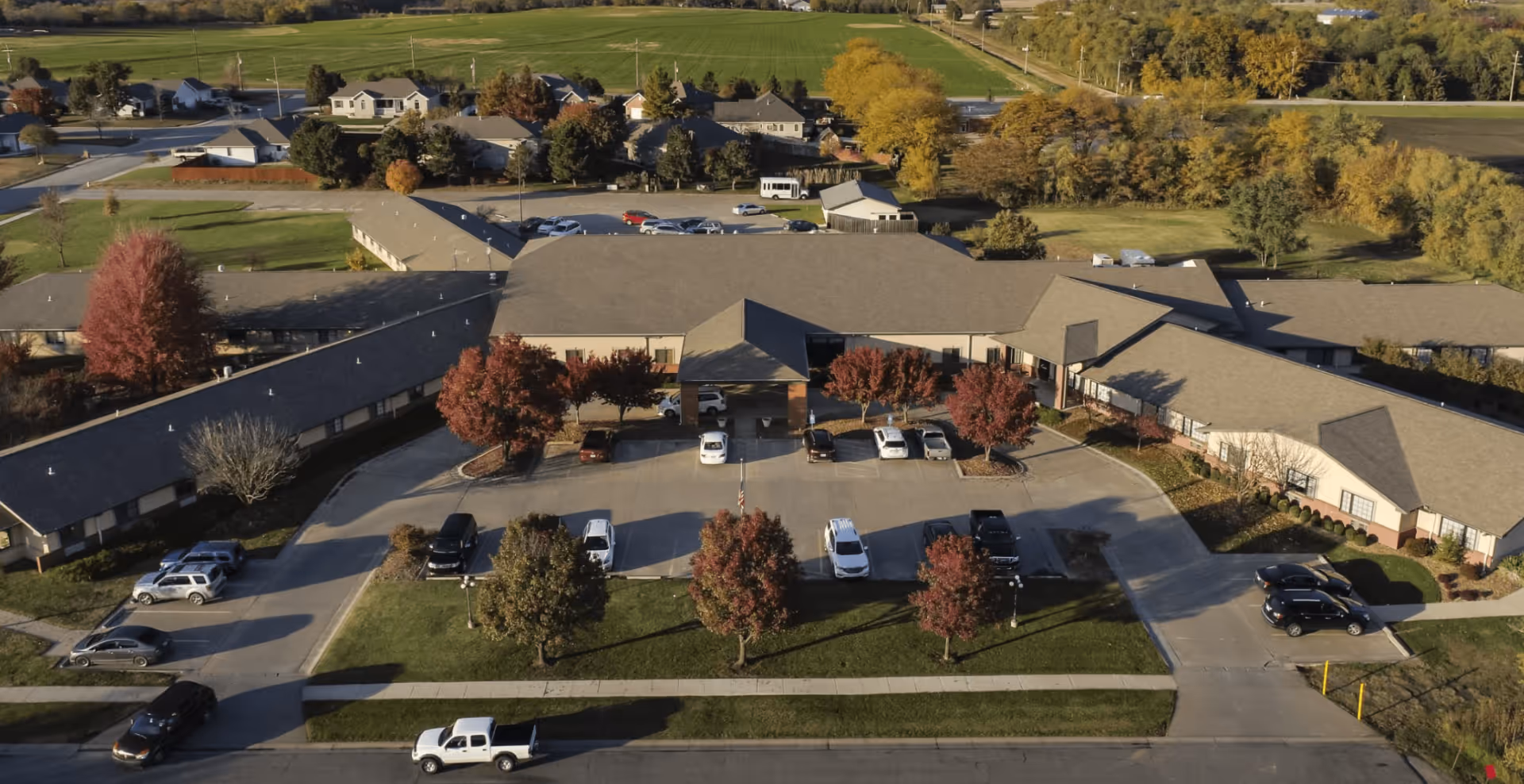 Aerial view of a single-story senior living facility showing the main entrance, parking lot, and surrounding trees and fields.