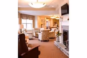 A cozy living room area in a retirement community featuring comfortable armchairs and sofas arranged around a stone fireplace. The room is warmly lit with ceiling lights and natural light coming through large windows with valances. A flat-screen TV is mounted above the fireplace, and a small table with a flower vase is placed nearby.