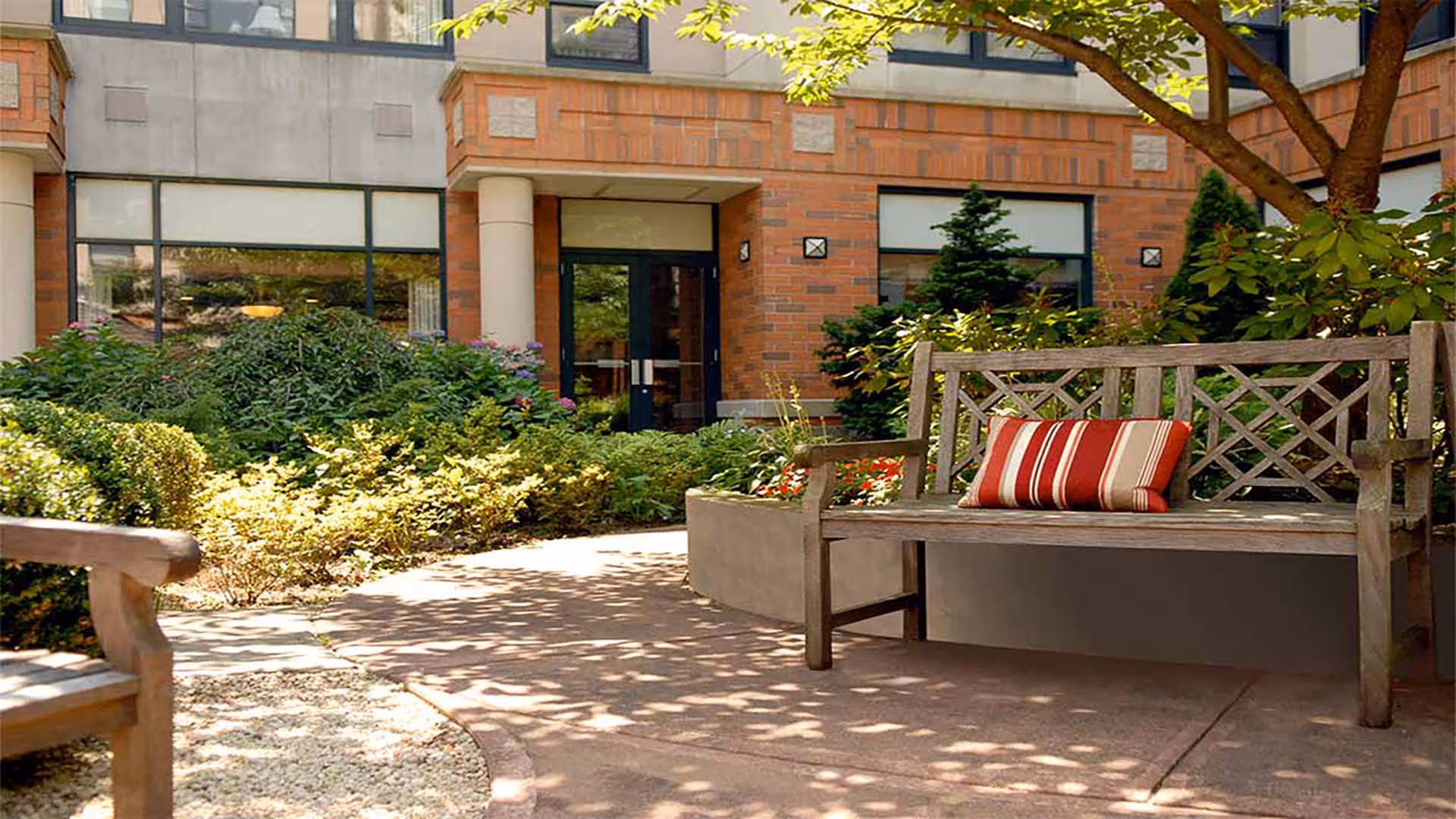 Outdoor seating area at Atria Cutter Mill featuring wooden benches with striped cushions, surrounded by greenery and plants, with the building's brick exterior and large windows in the background.