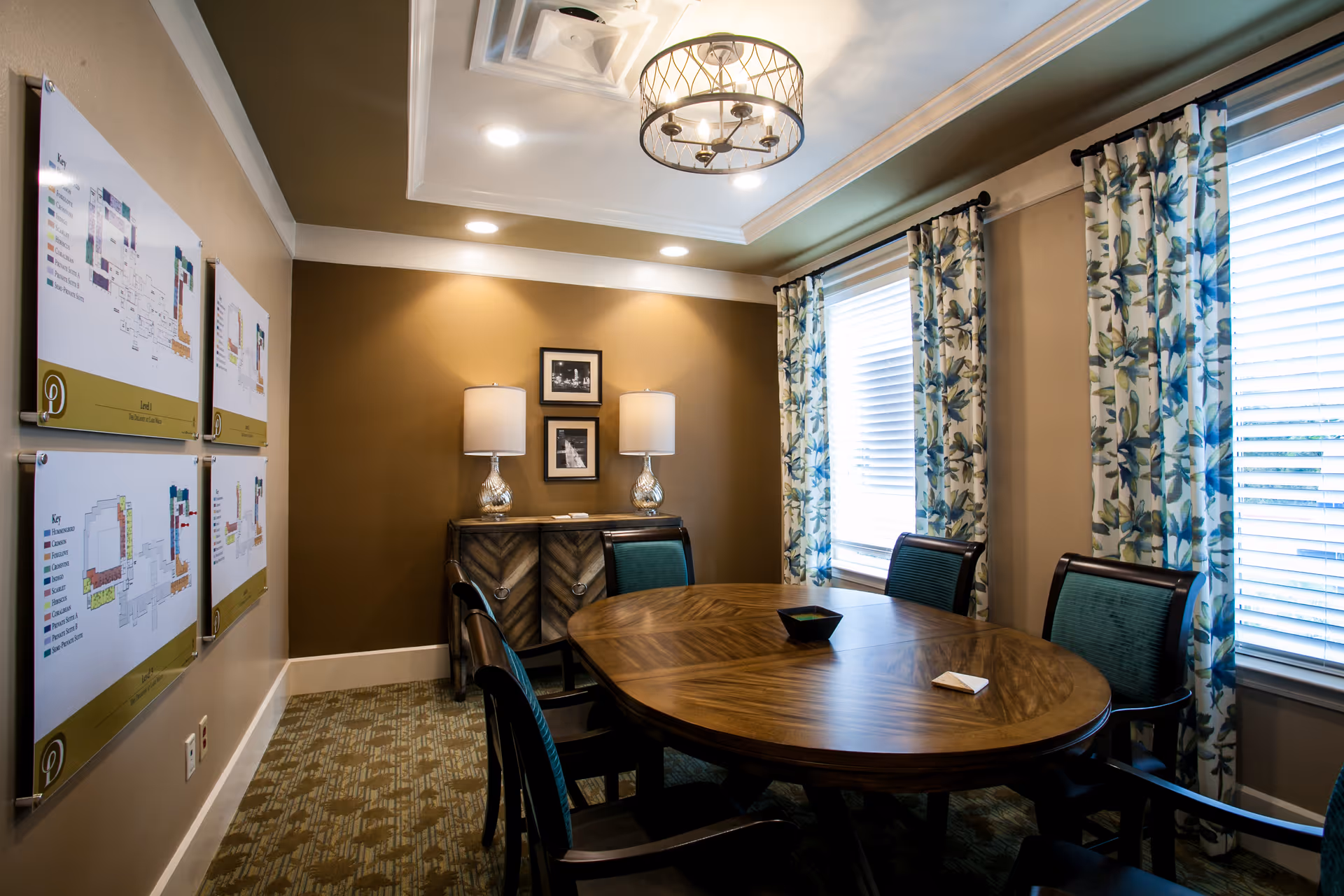 Round wooden table surrounded by upholstered chairs in a well-lit meeting room with floral curtains, wall-mounted maps, and table lamps.