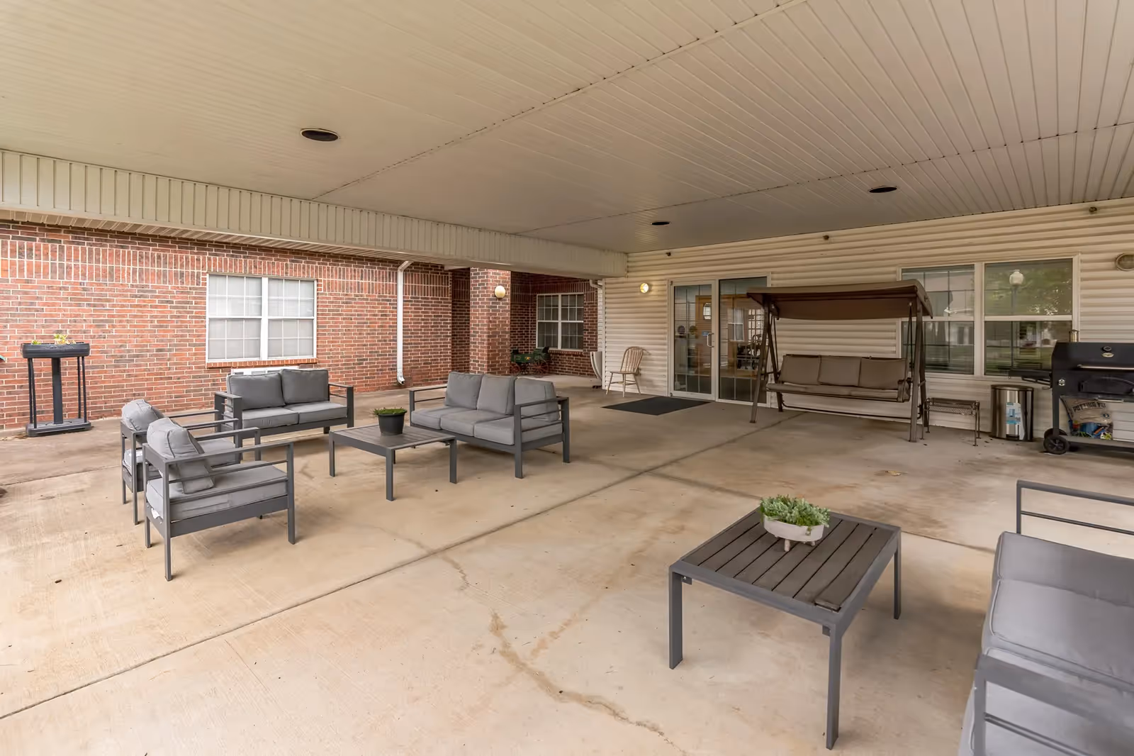 Covered outdoor patio area with modern gray cushioned seating including chairs, sofas, and a coffee table. There is a swing bench, a grill, and potted plants on the tables. The area is adjacent to a brick and siding building with windows and a glass door.