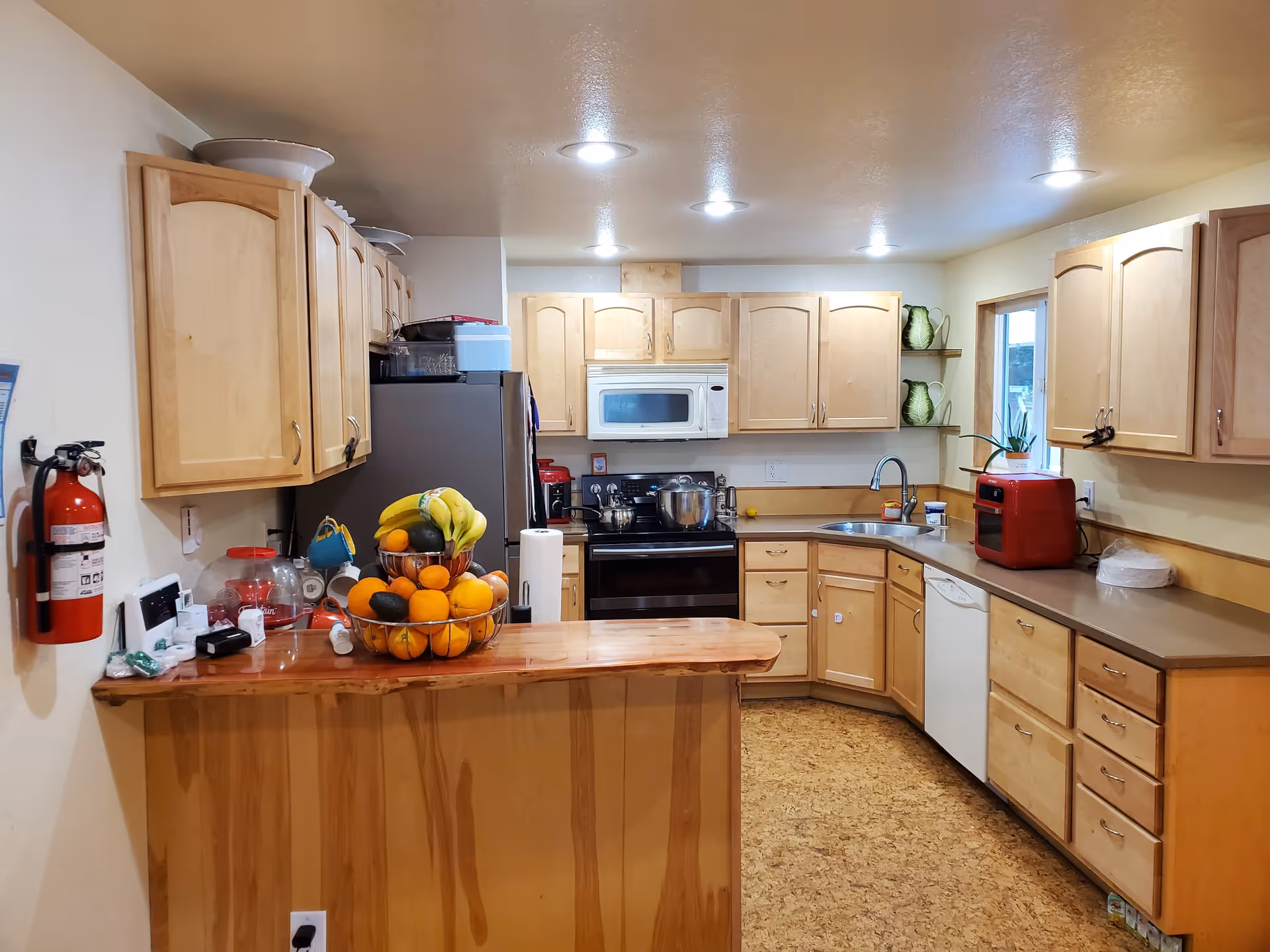 A well-lit kitchen with wooden cabinets and a wooden countertop island. The kitchen features a stove with a microwave above it, a refrigerator, a dishwasher, a sink, and various kitchen appliances including a red toaster oven. There is a tiered fruit basket with bananas, oranges, and avocados on the countertop island. A fire extinguisher is mounted on the wall to the left.