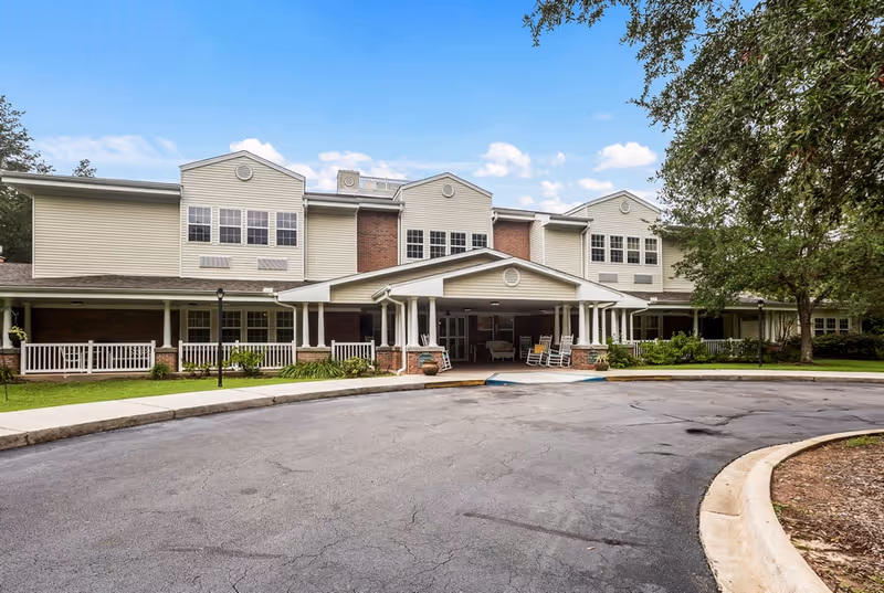 Front exterior of a two-story assisted living building with a covered circular drive, wraparound porch and rocking chairs.