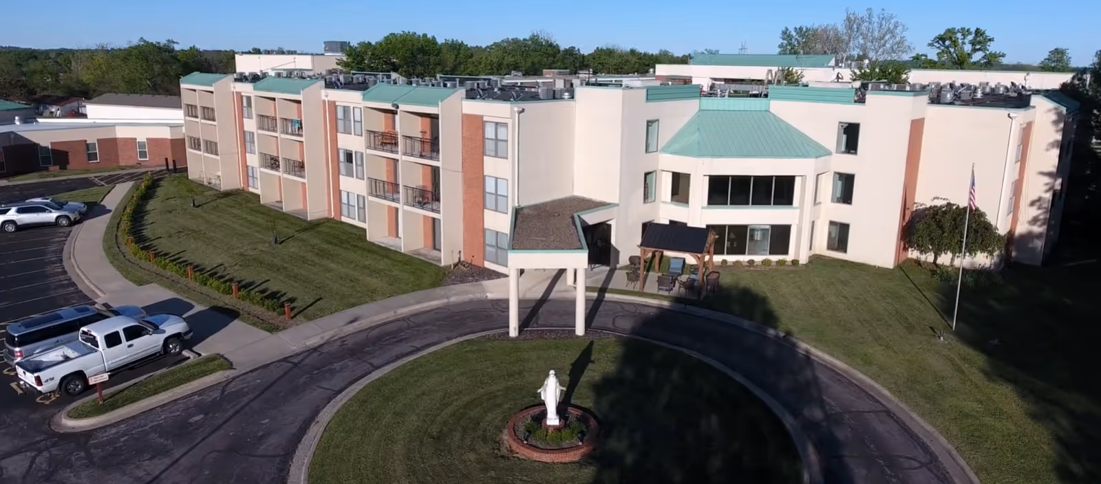 Aerial view of a multi-story senior living building with a circular driveway, front lawn, statue, and parked vehicles.
