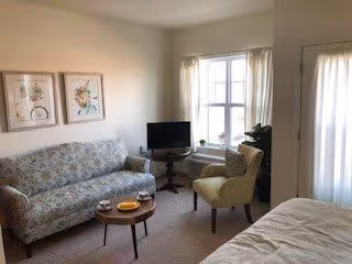 Studio living area with a floral sofa, armchair, small TV, coffee tables, and a bed by a curtained window.