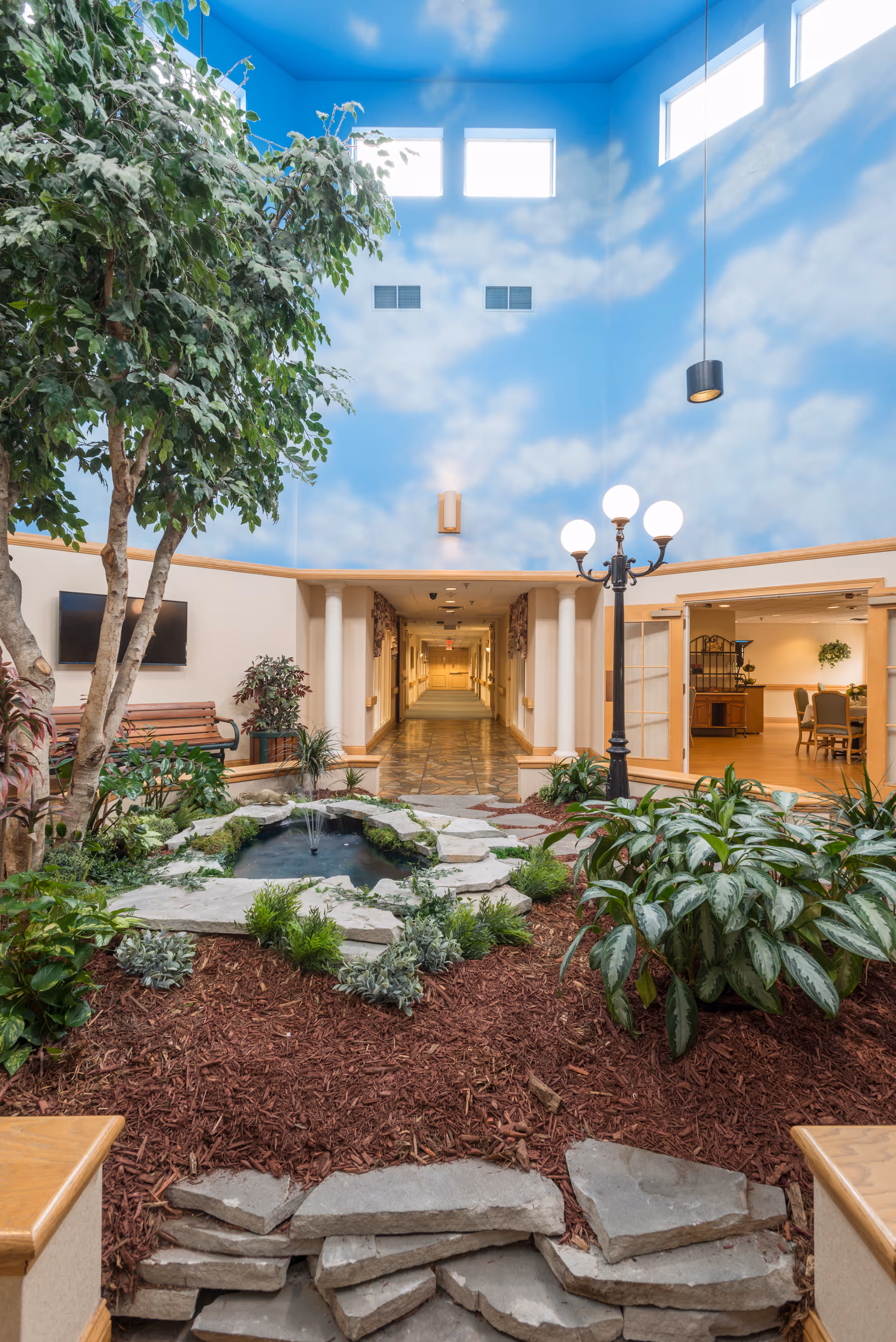 Indoor garden area with a small pond and fountain surrounded by plants and mulch, a tall tree on the left, a bench, and a hallway leading further into the facility. The ceiling is painted blue with white clouds, and there are high windows letting in natural light. A black streetlamp-style light fixture with four globes is near the pond. To the right, there is an open room with dining tables and chairs.