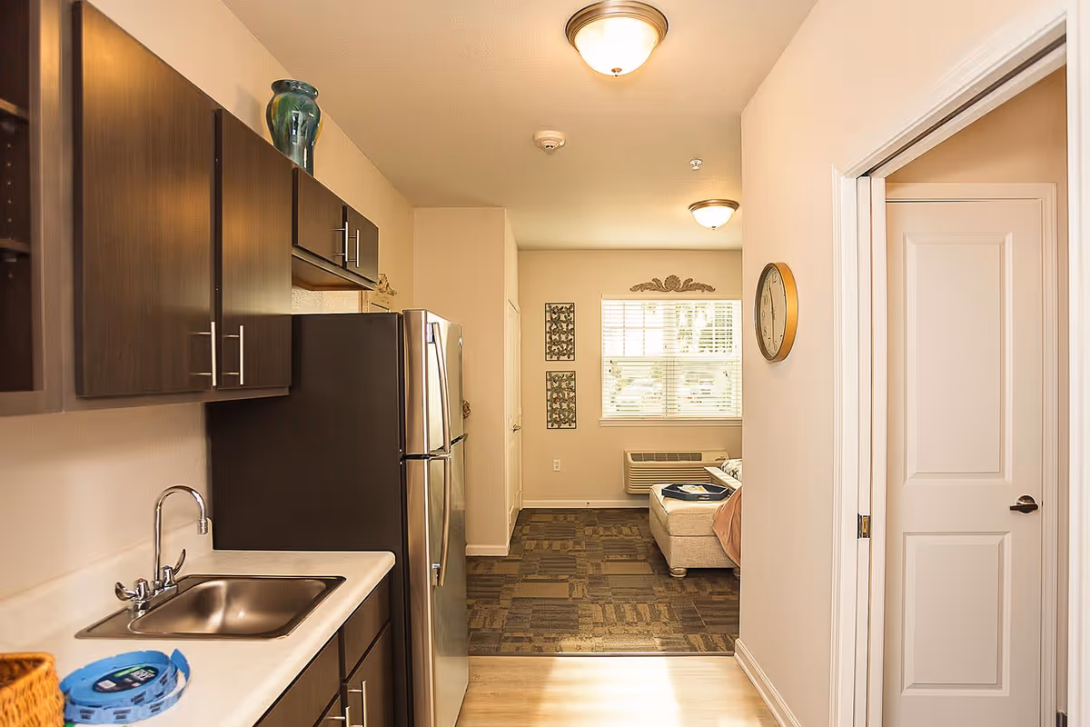 Interior view of a senior living facility apartment showing a small kitchen area with dark wood cabinets, a stainless steel refrigerator, and a sink. The kitchen opens into a living area with a window, wall decorations, and a beige ottoman with a tray on top. A clock is mounted on the wall near a white door.