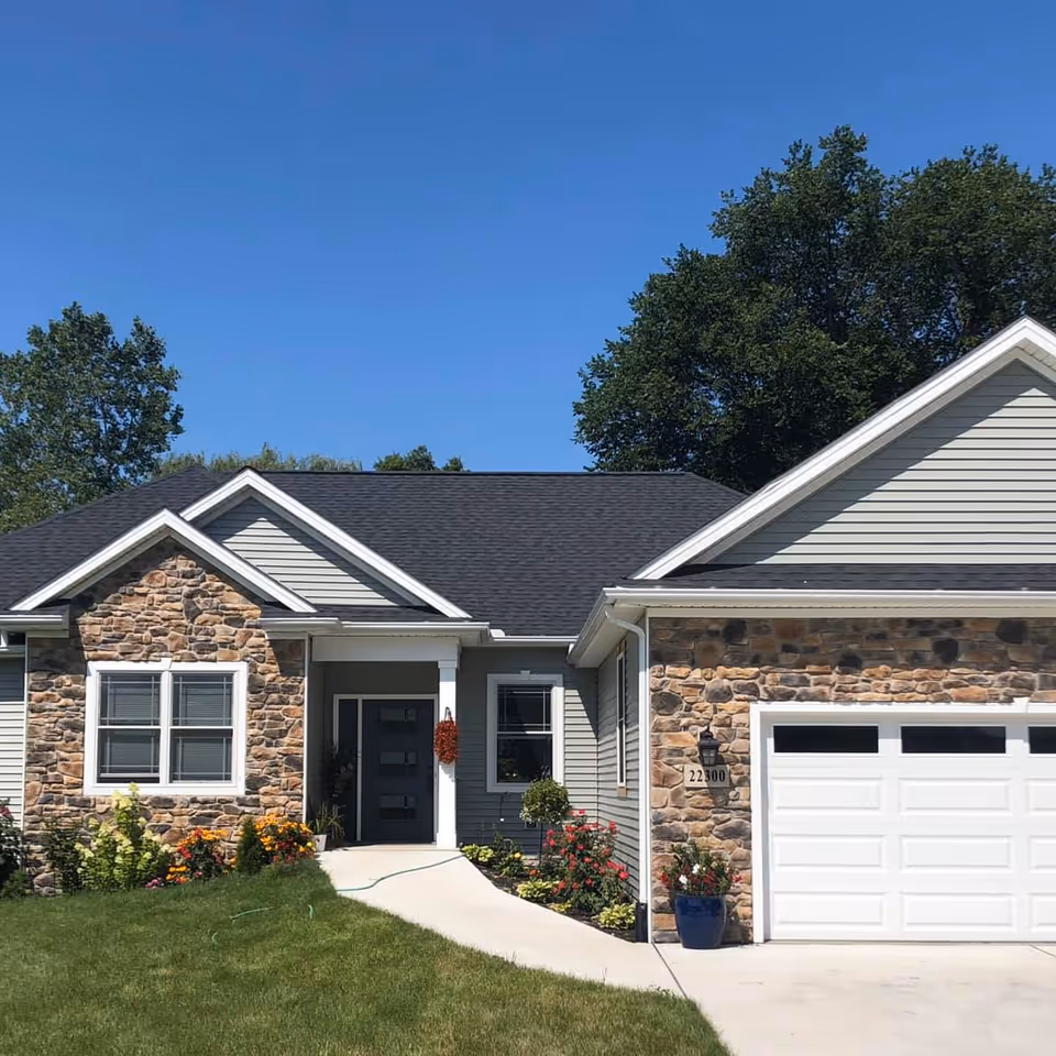 Front exterior view of a single-story senior living facility named Waltz Manor Senior Home with stone and siding facade, a black roof, a white garage door, a walkway leading to the front door, and landscaped flowers and shrubs.
