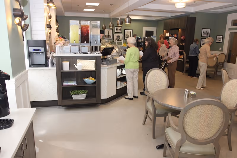 A group of elderly people standing in line at a beverage and snack counter in a well-lit dining area with round tables and upholstered chairs. The room has light green walls, framed pictures, and a modern, clean design.