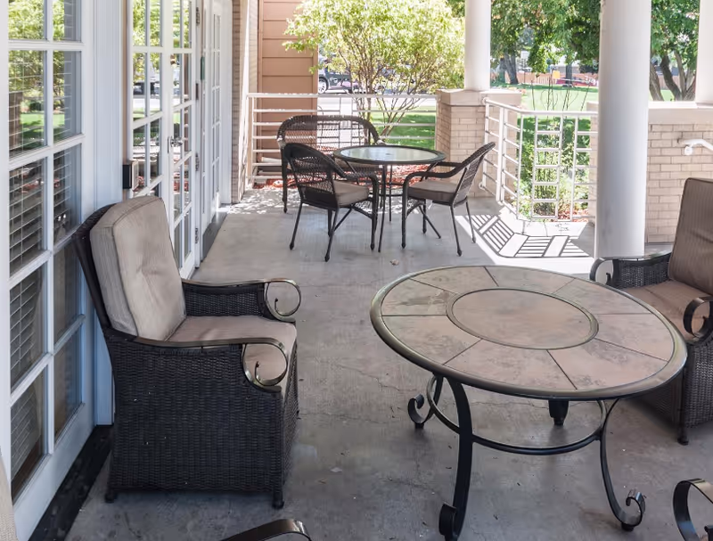 Outdoor covered patio area with cushioned wicker chairs and round tables. The patio is adjacent to a building with large glass doors and windows, overlooking a green lawn with trees and bushes.