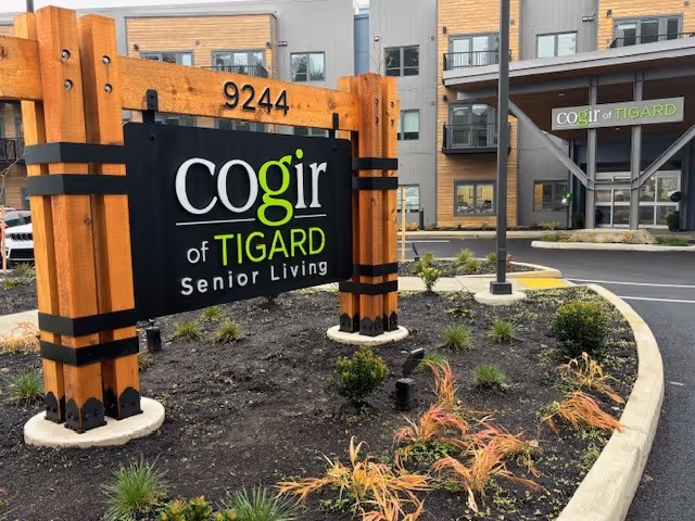 Outdoor view of the entrance area of Cogir of Tigard Senior Living facility, featuring a large wooden sign with the facility's name and address number 9244, surrounded by landscaping with small plants and shrubs, and the building entrance visible in the background.