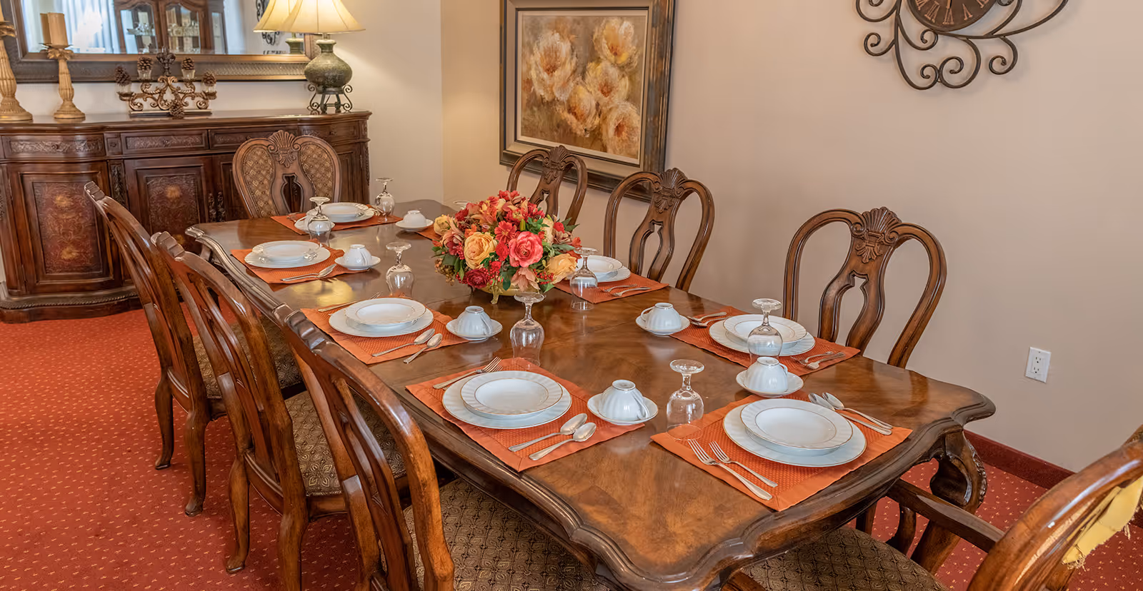 A formal dining room with a large wooden dining table set for eight people. The table has orange placemats, white plates, cups, silverware, and upside-down wine glasses. A floral centerpiece with red, orange, and yellow flowers is in the middle of the table. The room has a red carpet, a wooden sideboard with decorative items, a lamp, a large mirror, a floral painting on the wall, and a decorative wall clock.