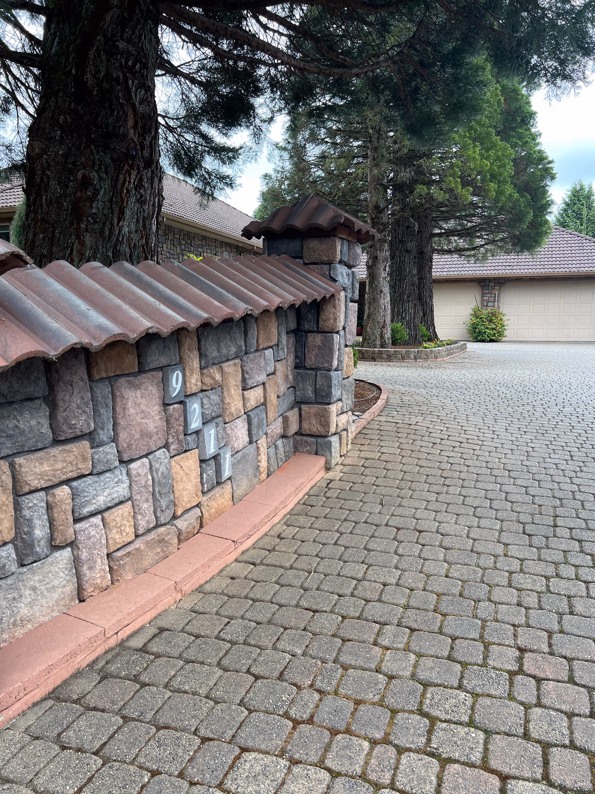 Stone boundary wall with address numbers beside a cobblestone driveway leading to a garage among large trees.