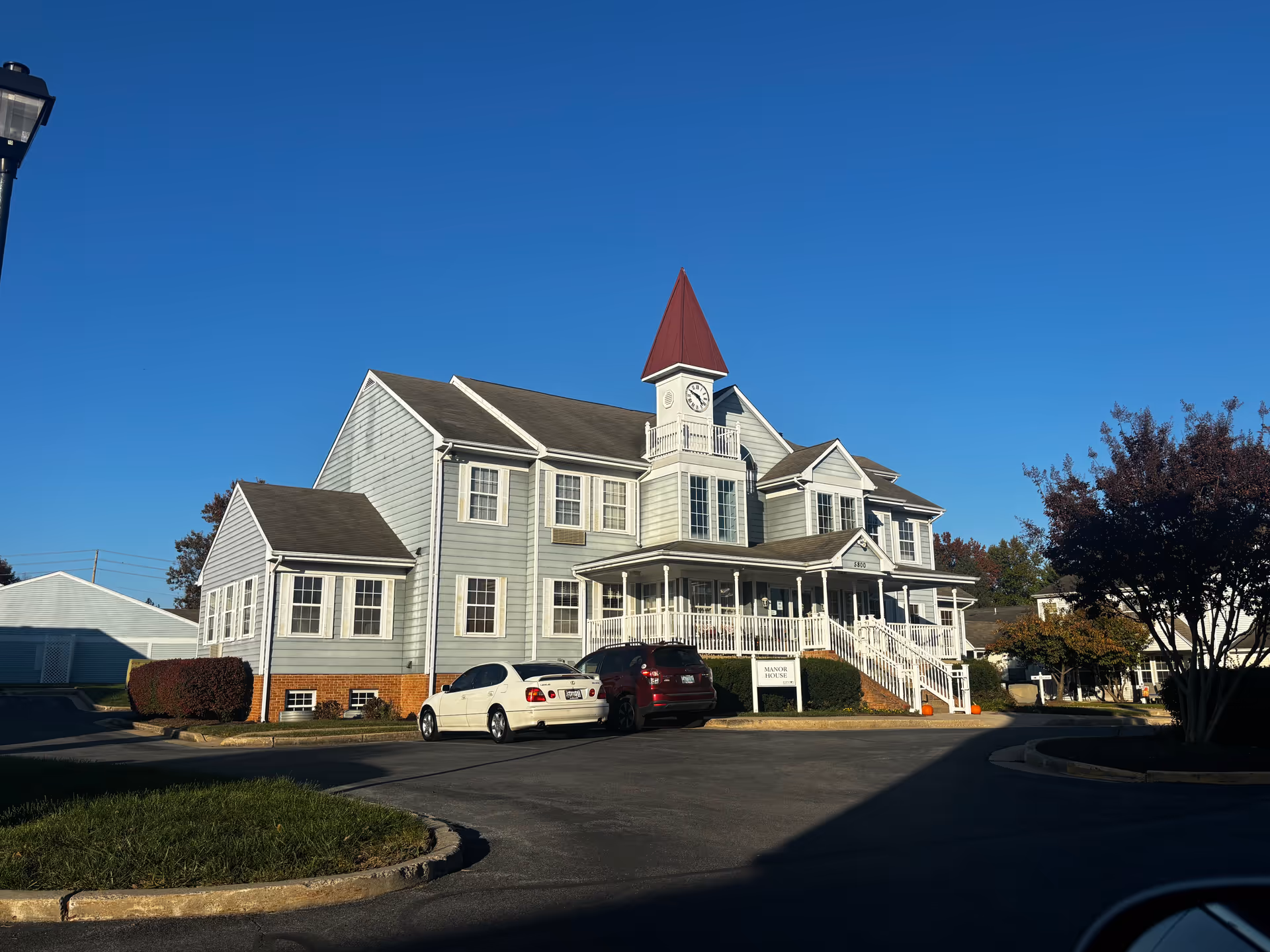 Exterior view of a two-story building with light gray siding and a red pointed clock tower on top. There is a white porch with railings and stairs leading up to the entrance. Two cars are parked in front of the building, and there are trees and bushes around the parking area under a clear blue sky.