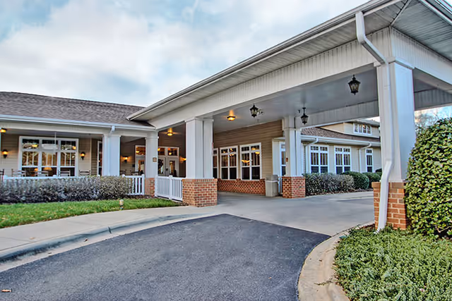 Entrance of a senior living facility with a covered driveway supported by white columns with brick bases, surrounded by well-maintained shrubs and greenery under a partly cloudy sky.
