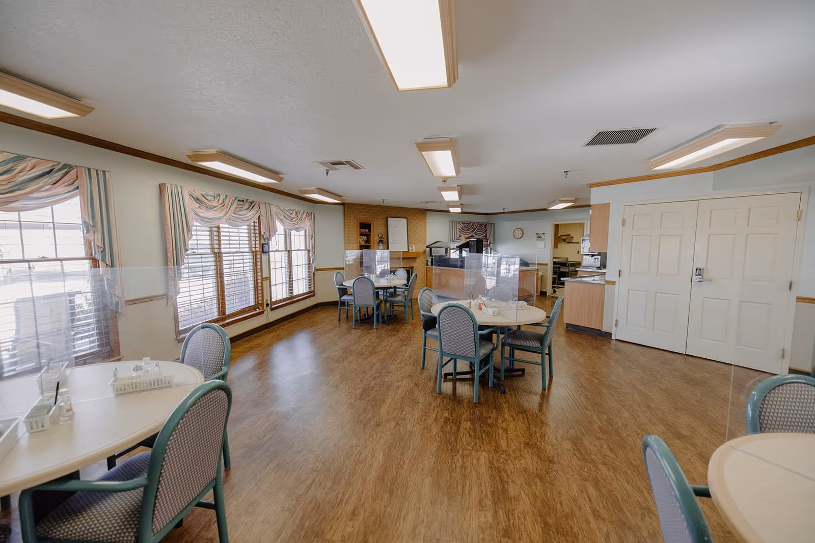 Interior view of the dining room at Redbud Plaza, featuring tables with chairs, large windows with curtains, and a warm wooden floor.