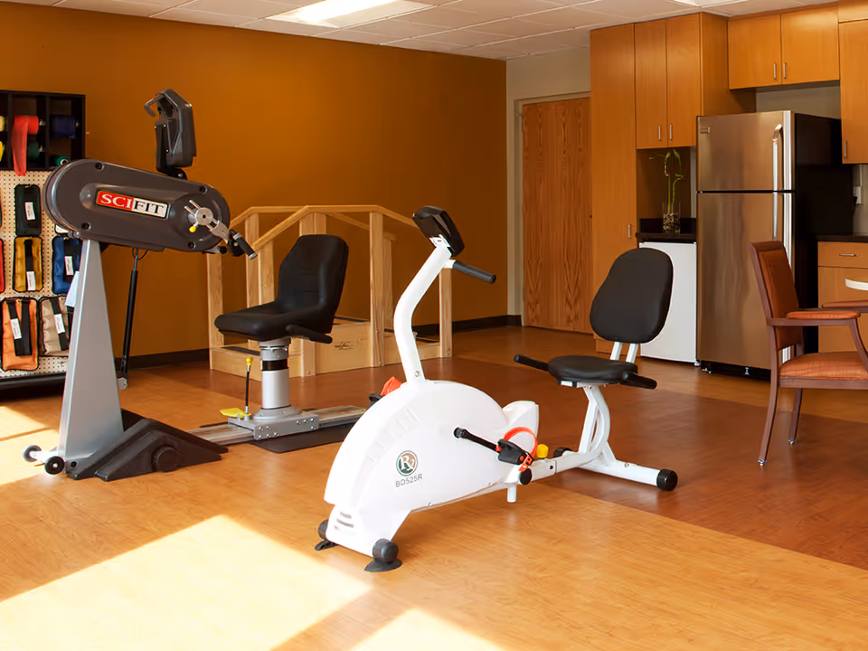 A room with exercise equipment including a recumbent bike and a seated arm exercise machine. The room has wooden flooring, a brown wall, and a kitchenette area with a refrigerator, cabinets, and chairs.