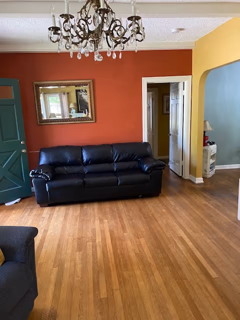 Living room with hardwood floors, a black leather sofa against a red accent wall, a chandelier overhead, and colored doors and archways leading to adjacent rooms.