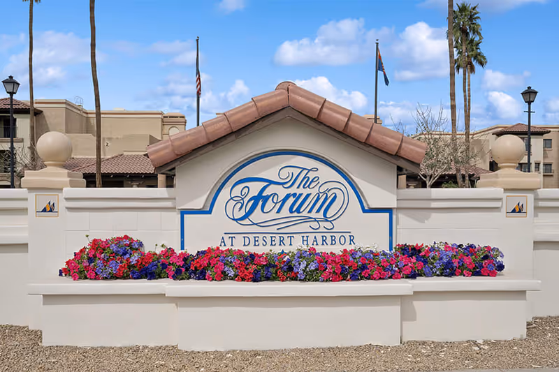 Entrance sign for The Forum at Desert Harbor with colorful flowers in front, beige walls, and buildings in the background under a blue sky with some clouds.