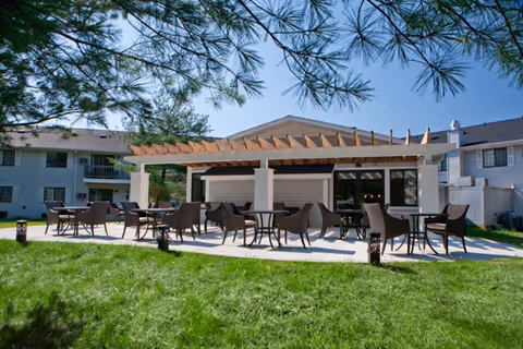 Outdoor patio area with multiple tables and chairs under a wooden pergola, surrounded by green grass and trees, with residential buildings in the background.
