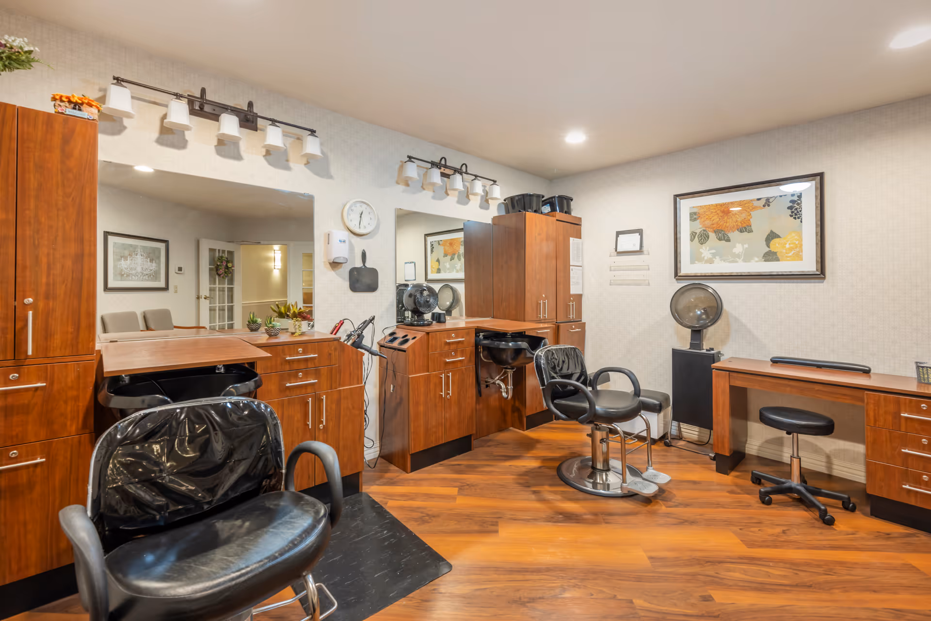 Interior view of a hair salon area with wooden cabinets, two black salon chairs, a hair washing station, a desk with a black rolling stool, a wall clock, and framed floral artwork on the wall.