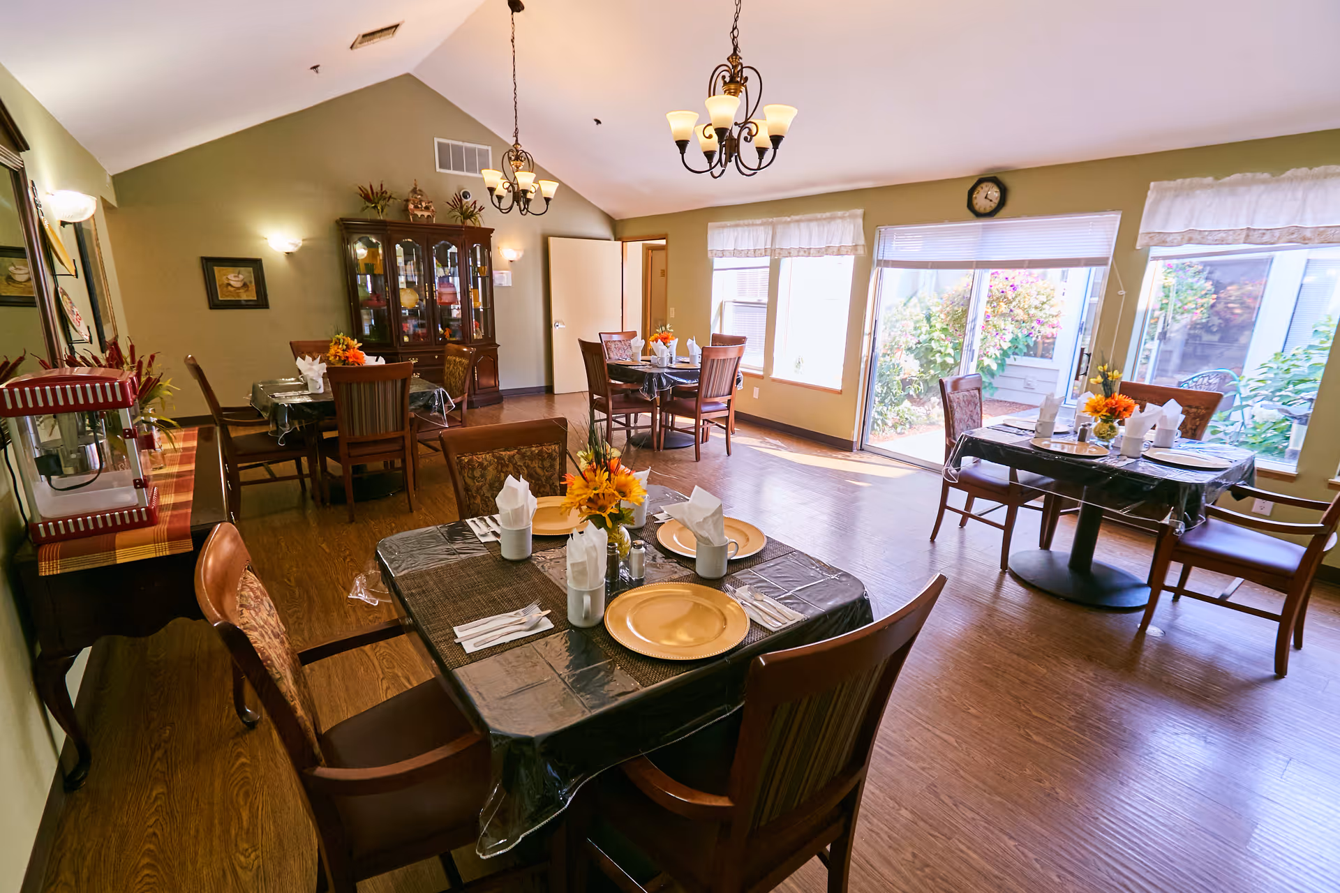 A bright dining room with multiple tables set for four people each, featuring yellow plates, white napkins, and floral centerpieces. The room has wooden floors, green walls, two chandeliers, a china cabinet, and large windows with a view of a garden outside.