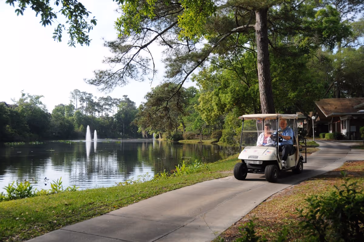 Two elderly people riding a golf cart on a paved path next to a lake with fountains, surrounded by lush green trees and a building in the background.
