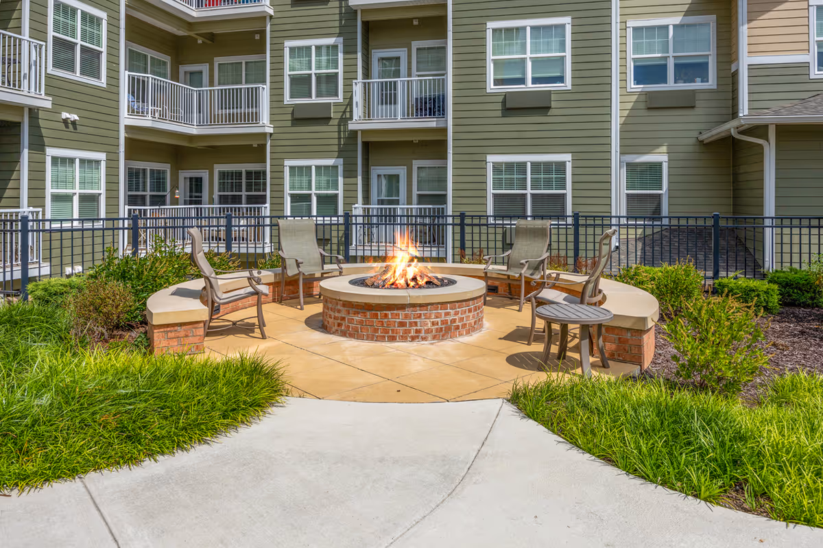 Outdoor seating area with a circular brick fire pit surrounded by chairs and curved benches, set in front of a green multi-story residential building with balconies and windows.