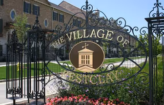 Ornate wrought-iron entrance gate with a circular emblem reading "The Village of Westcliff" in front of a residential building and landscaping.