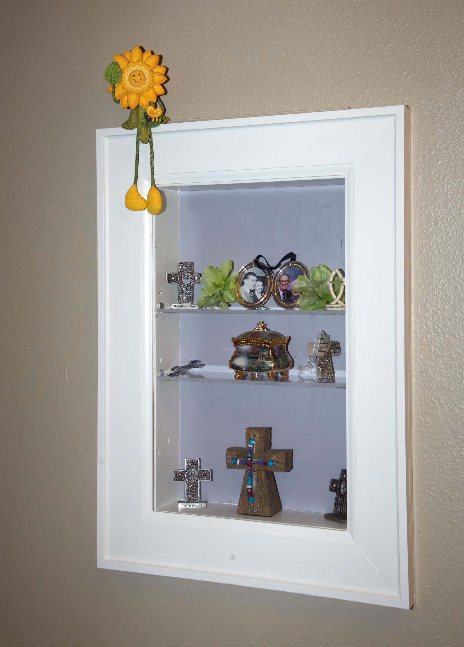 Wall-mounted white display cabinet holding small crosses, framed photos and decorative trinkets with a hanging sunflower ornament above.