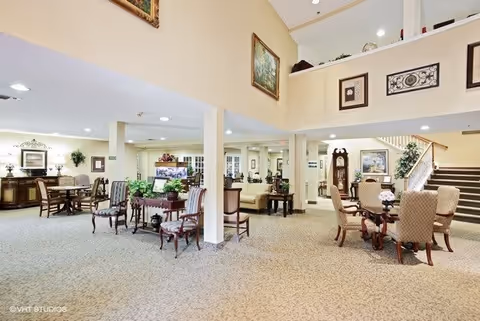 A spacious lobby area with elegant furnishings, including dining tables and chairs, decorative plants, and artwork on the walls. The space features a staircase leading to an upper level.